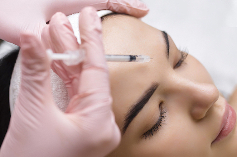 Woman with eyes closed receiving injection with needle between eyebrows with gloved hand.